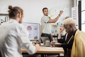 Um Homem Africano Aponta Para Um Quadro De Vidro Exibindo Um Relatório Financeiro Enquanto Discute Com Três Parceiros Homens Multiraciais Em Um Ambiente De Escritório Moderno.