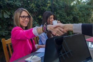 Duas Mulheres De Negócios Dando As Mãos Em Um Ambiente Iluminado, Com Plantas Verdes Ao Fundo, Simbolizando O Networking E A Colaboração.