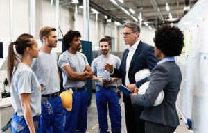 Um Gerente Corporativo Falando Para Um Grupo De Trabalhadores Manuais Durante Uma Reunião De Equipe Dentro De Uma Fábrica.