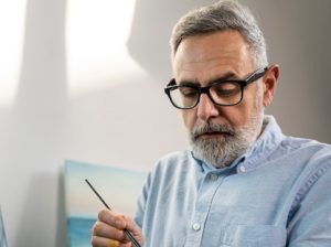 Um Plano Médio De Um Homem Concentrado Em Pintar Com Aquarelas, Cercado Por Materiais De Arte E Uma Tela.